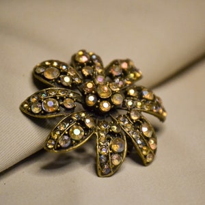 Puede incluir: Un broche dorado en forma de flor con pedrer&iacute;a iridiscente. El broche tiene un dise&ntilde;o detallado con m&uacute;ltiples p&eacute;talos y una piedra central.