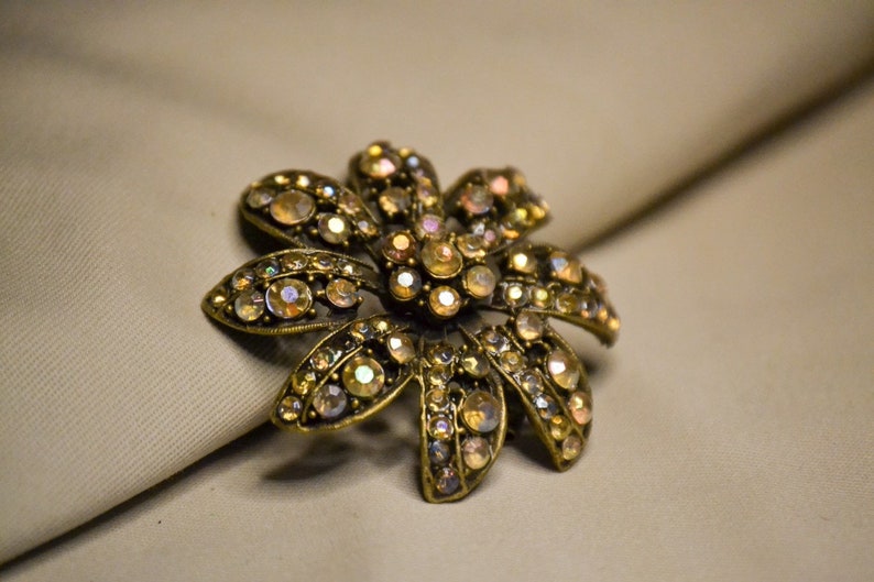 Puede incluir: Un broche dorado en forma de flor con pedrer&iacute;a iridiscente. El broche tiene un dise&ntilde;o detallado con m&uacute;ltiples p&eacute;talos y una piedra central.