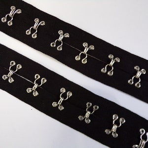 Puede incluir: Dos tiras de tela negra con cierres de gancho y ojo plateados. Las tiras están cosidas juntas con los cierres enfrentados.