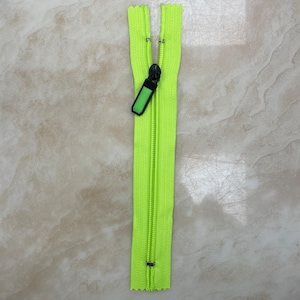 May include: A neon green zipper with a black pull tab and a green accent. The zipper is open, revealing the teeth. The zipper is made of a durable material and is suitable for various sewing projects.
