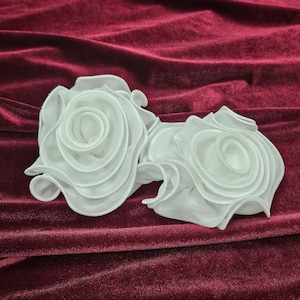 Può includere: Due fiori di tessuto bianco a forma di rosa su uno sfondo di velluto bordeaux.
