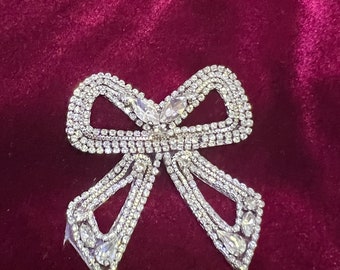 May include: A silver bow-shaped embellishment with rhinestones on a burgundy velvet background.