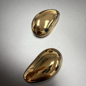 Puede incluir: Dos objetos decorativos dorados en forma de lágrima. Los objetos tienen una superficie lisa y reflectante, y están colocados sobre un fondo gris. Las formas son similares a una concha de mejillón o a una judía grande.