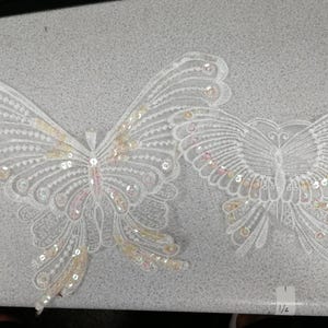 Könnte beinhalten: Zwei weiße schmetterlingsförmige Spitzenapplikationen mit aufwendiger Stickerei und irisierenden Pailletten. Der größere Schmetterling befindet sich links, der kleinere rechts. Ein kleines weißes Etikett mit dem Bruch 1/2 ist unten rechts sichtbar.