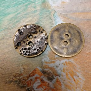 May include: Two antique brass buttons. One button has a textured, pitted surface with several holes. The other button is smooth with two holes for sewing. The buttons are round and have a vintage aesthetic.