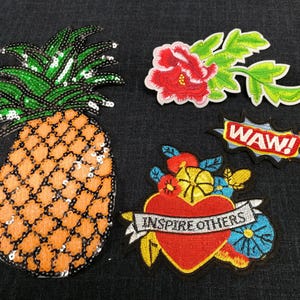 May include: A collection of embroidered patches. Includes a sequined pineapple, a red flower with green leaves, a heart with the words "INSPIRE OTHERS", and a "WAW!" speech bubble. Ideal for customising clothing or accessories.