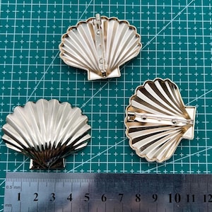 Op de afbeelding: Drie gouden metalen schelpvormige broches met een pin sluiting. De broches liggen op een groene grid achtergrond.