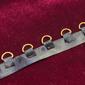 May include: A black fabric ribbon featuring a series of evenly spaced gold-toned metal D-rings attached with small black fabric loops. The ribbon has a smooth, slightly reflective texture and is positioned horizontally across the frame.