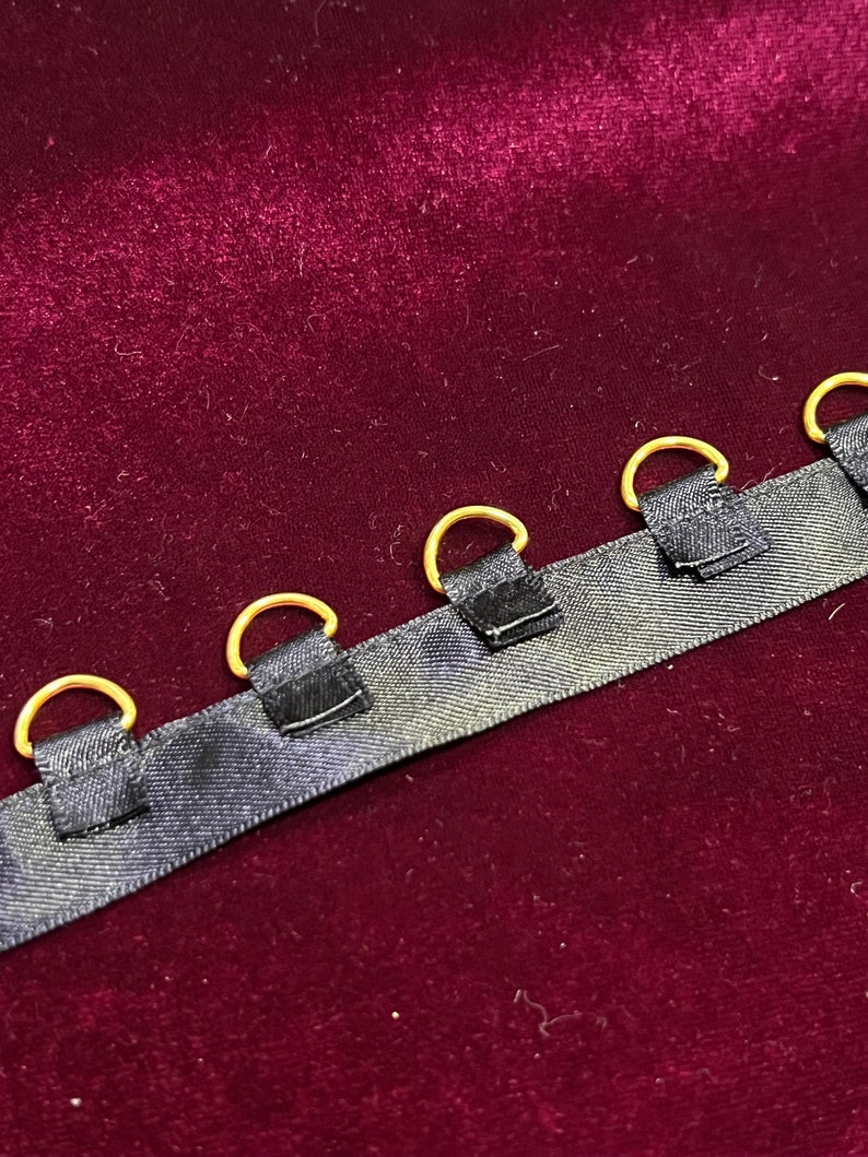 May include: A black fabric ribbon featuring a series of evenly spaced gold-toned metal D-rings attached with small black fabric loops. The ribbon has a smooth, slightly reflective texture and is positioned horizontally across the frame.