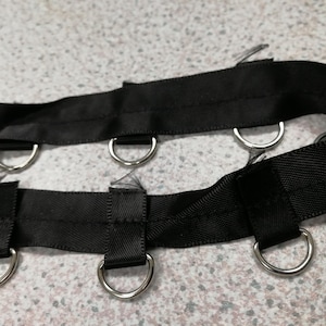 May include: A black fabric strap featuring two parallel rows of silver-toned metal D-rings attached with short fabric loops. The strap is constructed from a woven material with a smooth texture and visible stitching along the edges of the loops.