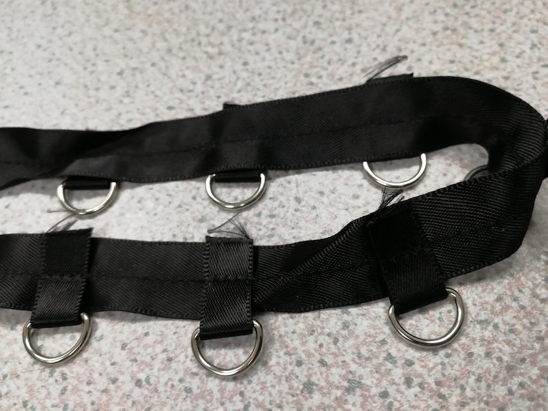 May include: A black fabric strap featuring two parallel rows of silver-toned metal D-rings attached with short fabric loops. The strap is constructed from a woven material with a smooth texture and visible stitching along the edges of the loops.