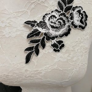 May include: Black and white floral embroidered applique. The applique features a large peony-like flower with detailed petals and leaves. The design is set against a white background, creating a striking contrast. Ideal for embellishing garments or accessories.