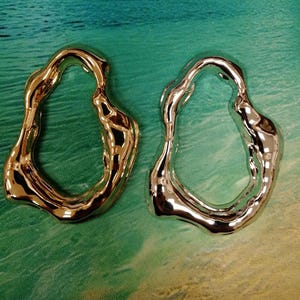 Könnte beinhalten: Zwei abstrakte, organisch geformte Metallobjekte. Eines ist goldfarben, das andere silberfarben. Beide haben eine glatte, reflektierende Oberfläche und eine unregelmäßige, offene Mitte. Der Hintergrund ist eine Mischung aus Türkis, Grün und Gelb.
