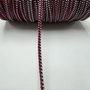 Puede incluir: Primer plano de un carrete de cordón trenzado rojo, blanco y negro. El cordón está enrollado firmemente alrededor del carrete y un tramo de cordón cuelga.
