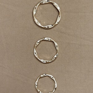 Op de afbeelding: Drie gouden, gedraaide metalen ringen in verschillende maten. De ringen zijn verticaal gerangschikt, met de grootste ring bovenaan en de kleinste ring onderaan.