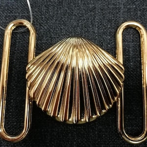 May include: A gold-coloured metal clasp featuring a seashell design in the centre. The clasp has two rectangular loops either side of the shell. The background is a dark grey fabric.