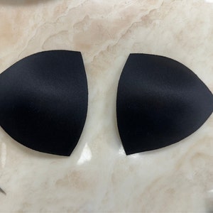 Könnte beinhalten: Zwei schwarze BH-Cups aus Schaumstoff. Die Cups sind dreieckig geformt und haben eine glatte, leicht glänzende Oberfläche.