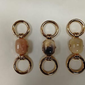 Könnte beinhalten: Drei dekorative Verbinder mit goldfarbenen Ringen und rechteckigen, perlenartigen Mittelstücken. Die Mittelstücke sind in den Farbtönen Pfirsich, Grau und Creme gehalten. Diese Verbinder sind wahrscheinlich für Schmuck oder Accessoires.