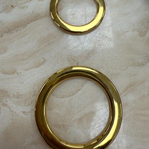 Könnte beinhalten: Zwei goldfarbene Metallringe in verschiedenen Größen. Die Ringe sind kreisförmig mit einer glatten, glänzenden Oberfläche. Der größere Ring befindet sich unten, der kleinere Ring darüber. Der Hintergrund ist eine helle Oberfläche mit einem dezenten Muster.