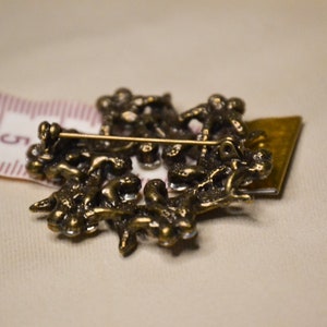 May include: A gold-toned brooch with a detailed design of intertwined branches and leaves. The brooch has a pin back and is decorated with clear rhinestones.