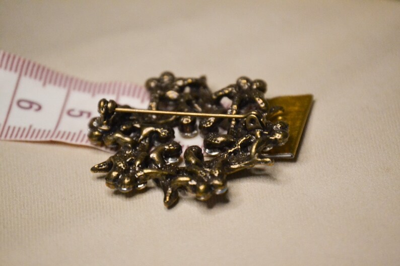 May include: A gold-toned brooch with a detailed design of intertwined branches and leaves. The brooch has a pin back and is decorated with clear rhinestones.