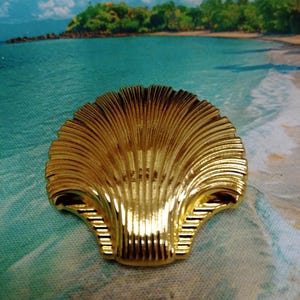Puede incluir: Un objeto decorativo dorado en forma de concha. El objeto tiene una superficie detallada y acanalada y una forma curva, que se asemeja a una concha de vieira. El fondo presenta una escena de playa con agua azul y una isla tropical.
