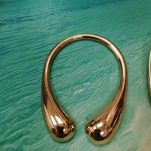 May include: A polished gold-tone metal accessory featuring a smooth, continuous U-shaped design with two rounded, teardrop-like ends that curve inward towards each other, creating an open, circular silhouette with a highly reflective, mirror-like finish.