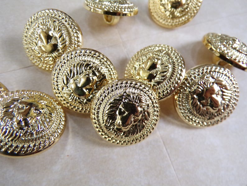 Puede incluir: Un grupo de ocho botones de metal dorado con un dise&ntilde;o de cabeza de le&oacute;n. Los botones tienen un dise&ntilde;o en relieve texturizado y un acabado brillante.