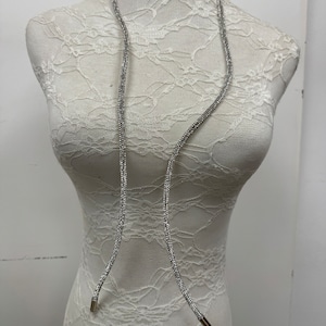 May include: A silver rhinestone chain necklace with a long, dangling design. The necklace is displayed on a white lace-covered mannequin.