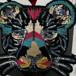 May include: A black embroidered patch in the shape of a tiger's face, embellished with sequins in gold, blue, and iridescent colors. The tiger's face has a geometric design with pink and turquoise accents.