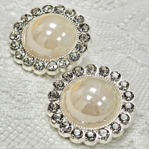 May include: Two round, silver-toned, decorative buttons with a white pearl-like center surrounded by clear rhinestones.