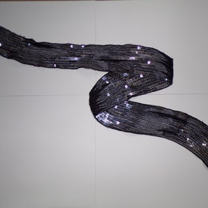 Könnte beinhalten: Ein schwarzer Pailetten-Schal mit silbernem Schimmer. Der Schal ist gefaltet und auf einer weißen Oberfläche ausgelegt.