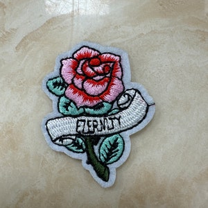 May include: Embroidered iron-on patch featuring a pink and red rose with green leaves and a white banner that reads "Eternity".