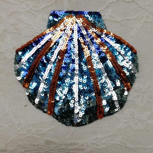 May include: A seashell-shaped patch embellished with sequins in shades of blue, silver, and copper. The design features radiating lines of copper and silver sequins against a background of dark blue and teal sequins, creating a textured effect.