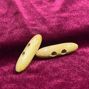 Puede incluir: Dos botones de madera con dos agujeros cada uno. Los botones son de color marrón claro y tienen un acabado liso y pulido.