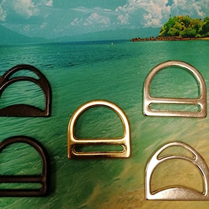 May include: An assortment of D-rings in various metallic finishes, including gold, silver, and black. The hardware is arranged against a backdrop of a tropical beach scene, showcasing the rings' shapes and colors.