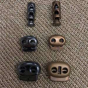 Può includere: Sei fermagli per cordino a levetta, tre neri e tre bronzo, con due fori ciascuno. I fermagli per cordino sono su uno sfondo a rete dorata.