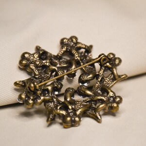 May include: A gold-toned brooch with a detailed design of intertwined branches and leaves. The brooch is adorned with clear rhinestones.