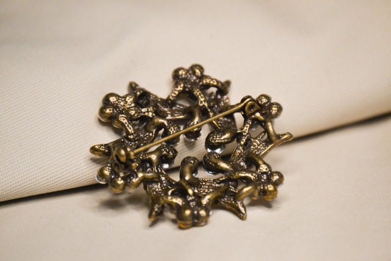May include: A gold-toned brooch with a detailed design of intertwined branches and leaves. The brooch is adorned with clear rhinestones.