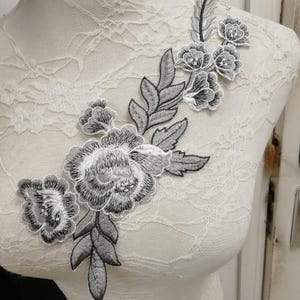 Könnte beinhalten: Grau-weiße, floral bestickte Applikation. Die Applikation zeigt detaillierte Stickereien von Blumen und Blättern. Das Design wird auf einer weißen Schaufensterpuppe präsentiert und zeigt die filigrane Handwerkskunst und die potenzielle Verwendung zur Verzierung von Kleidungsstücken oder Bastelarbeiten.