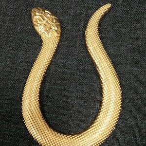May include: A gold-coloured snake-shaped brooch. The snake's head has a detailed pattern, and the body is textured with a dot pattern. The brooch is set against a dark background, highlighting its metallic sheen. A unique jewellery piece.