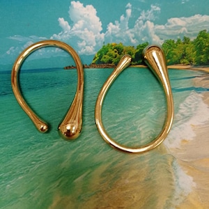 Peut inclure: Deux bracelets dorés ouverts avec un design en forme de goutte d'eau. Les bracelets sont présentés sur un fond coloré avec une scène de plage. Les bijoux ont une surface brillante et réfléchissante.