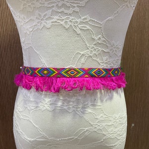 May include: A pink fringed belt with a colourful geometric pattern. The belt is made of woven fabric and has a tassel fringe.