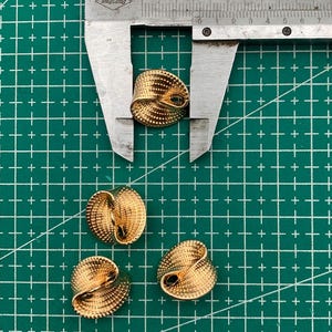 Op de afbeelding: Vier gouden metalen knopen met een swirl-ontwerp. De knopen worden gemeten met een liniaal.