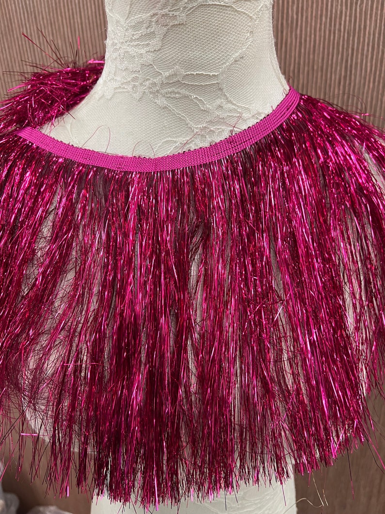 May include: A close-up of a pink metallic fringe trim. The fringe is made of thin, shiny strands of fabric.