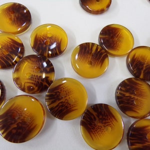 以下が含まれることがあります： 木目模様の茶色と黄色の丸いボタンが15個あります。ボタンには穴が2つあります。