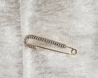 May include: A gold safety pin with a clear rhinestone design.