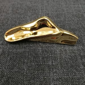 Op de afbeelding: Een goudkleurige, abstract gevormde broche met een glad, reflecterend oppervlak. De broche heeft een gebogen, organische vorm, die lijkt op een gestileerd landschap of een gesmolten object. De achtergrond is een donkergrijze stof.