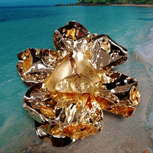 Puede incluir: Broche dorado en forma de flor con un acabado metálico texturizado. El broche tiene múltiples pétalos y un elemento central triangular. El fondo presenta una escena de playa con agua azul y una isla verde.