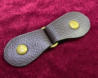 May include: A brown leather toggle clasp with two brass button closures. The clasp is designed to be used on a garment or bag.